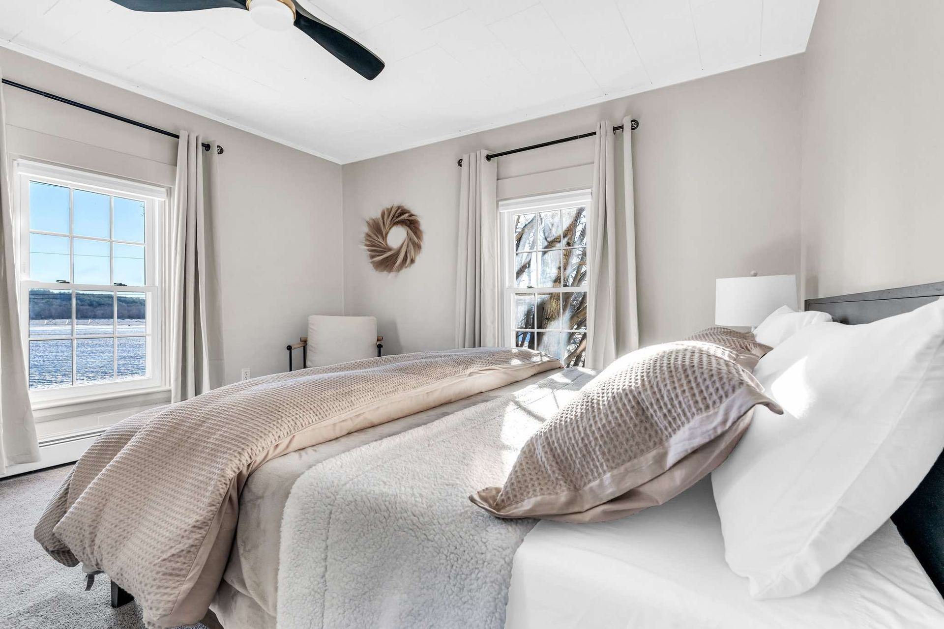 Cozy bedroom with bed, windows overlooking water, neutral tones, and a decorative wreath.
