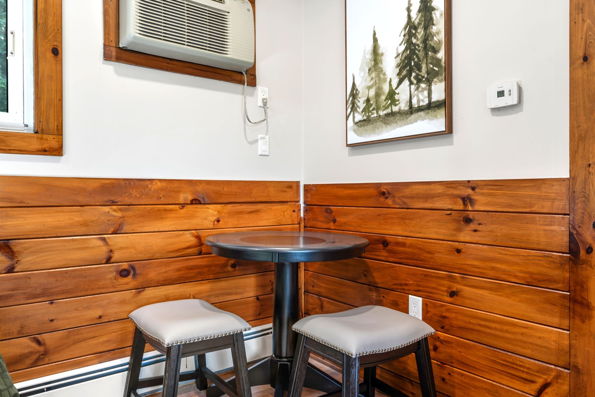 Corner dining area with wooden paneling, small table, two stools, AC unit, and framed artwork.