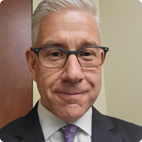 Man with gray hair and glasses wearing a suit, smiling slightly, in an office setting.