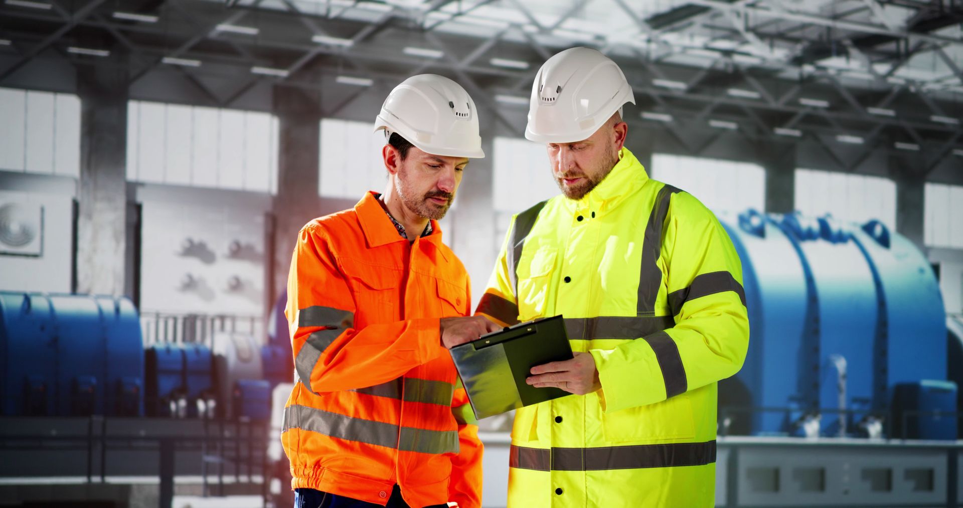 Two construction workers are looking at a clipboard in a factory.