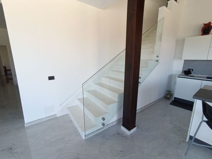 Un'area salotto esterna con patio, racchiusa da una struttura in legno con ampie vetrate, vista da dietro una recinzione metallica.