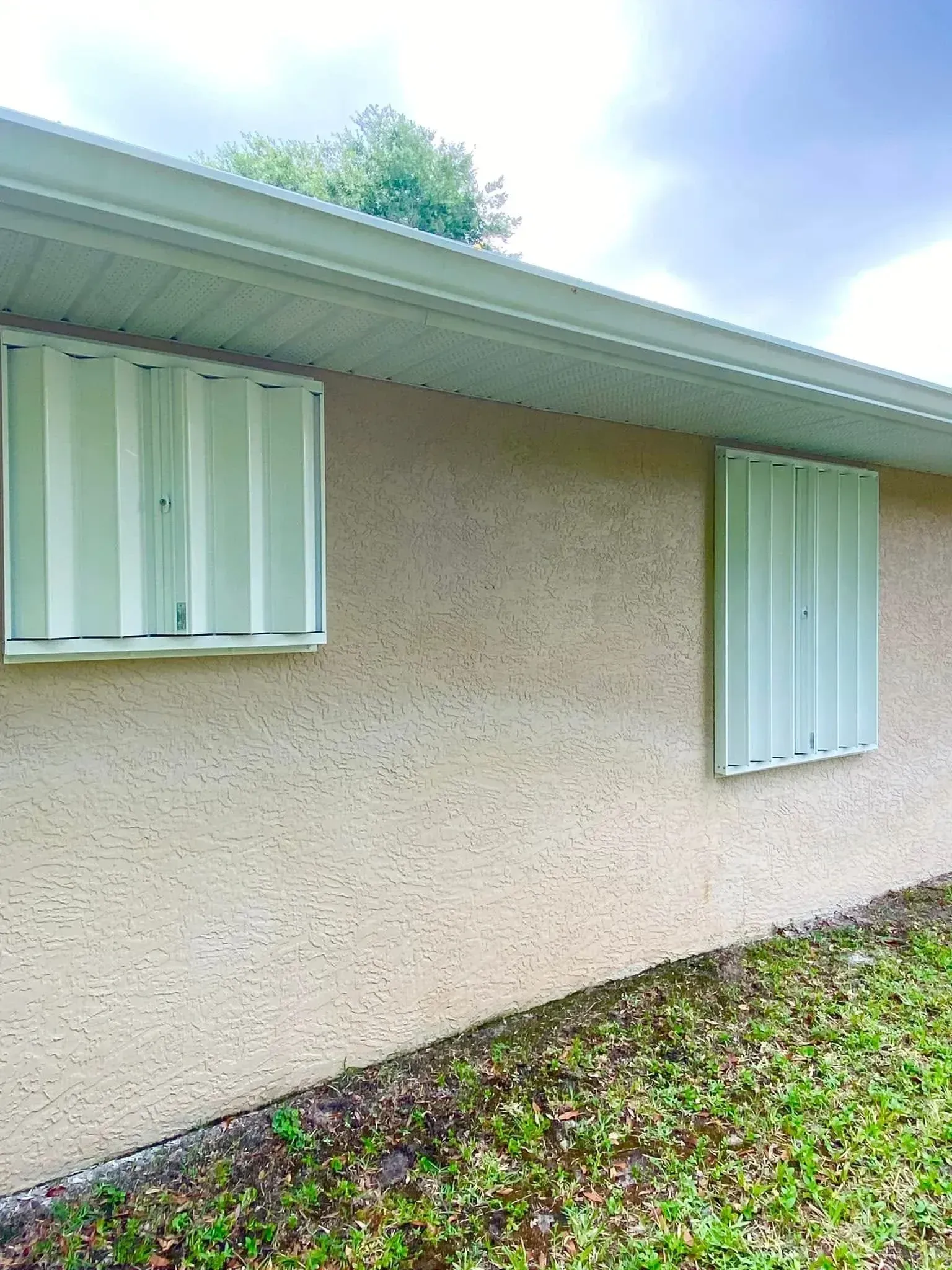 Exterior of a beige stucco house with two windows and white shutters under a sloped roof.