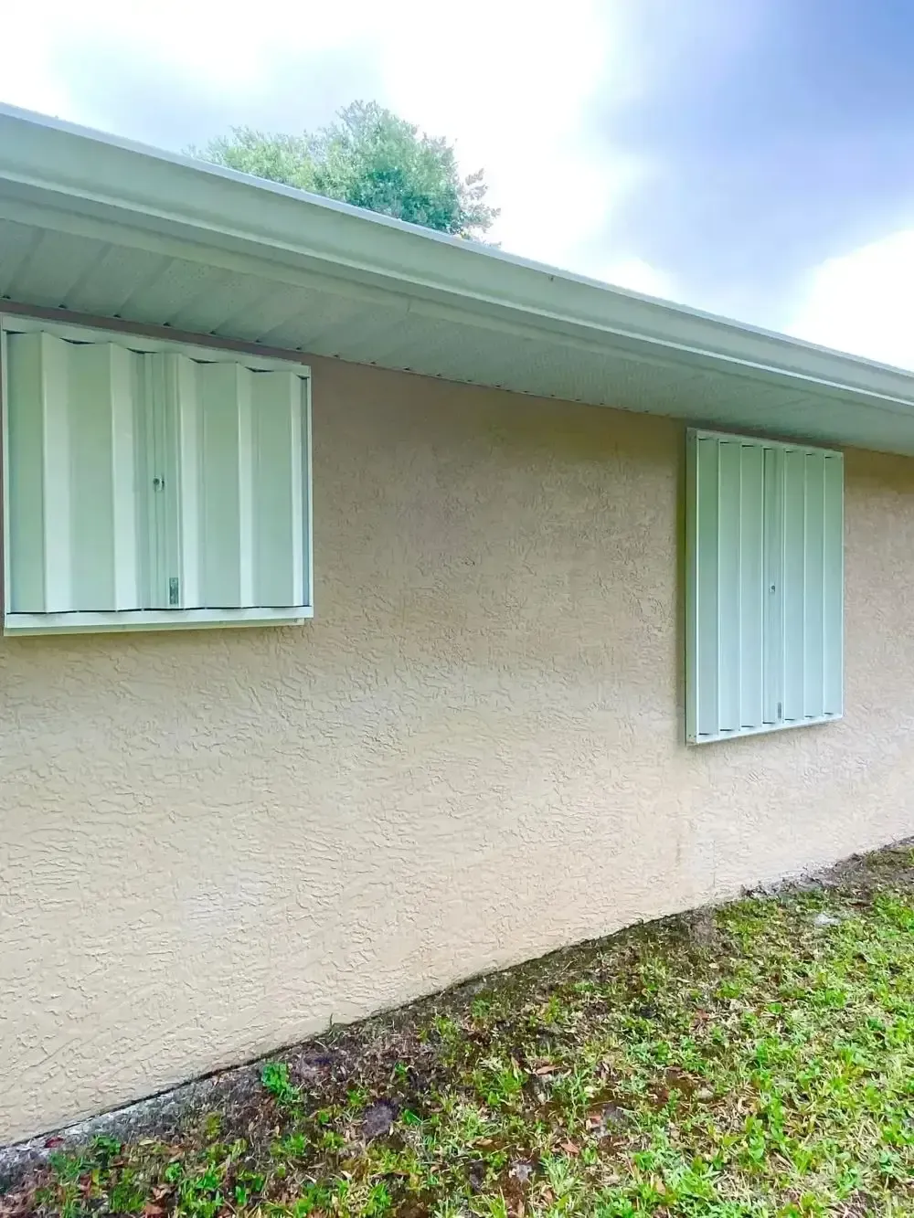 Exterior of a beige stucco house with two windows and white shutters under a sloped roof.
