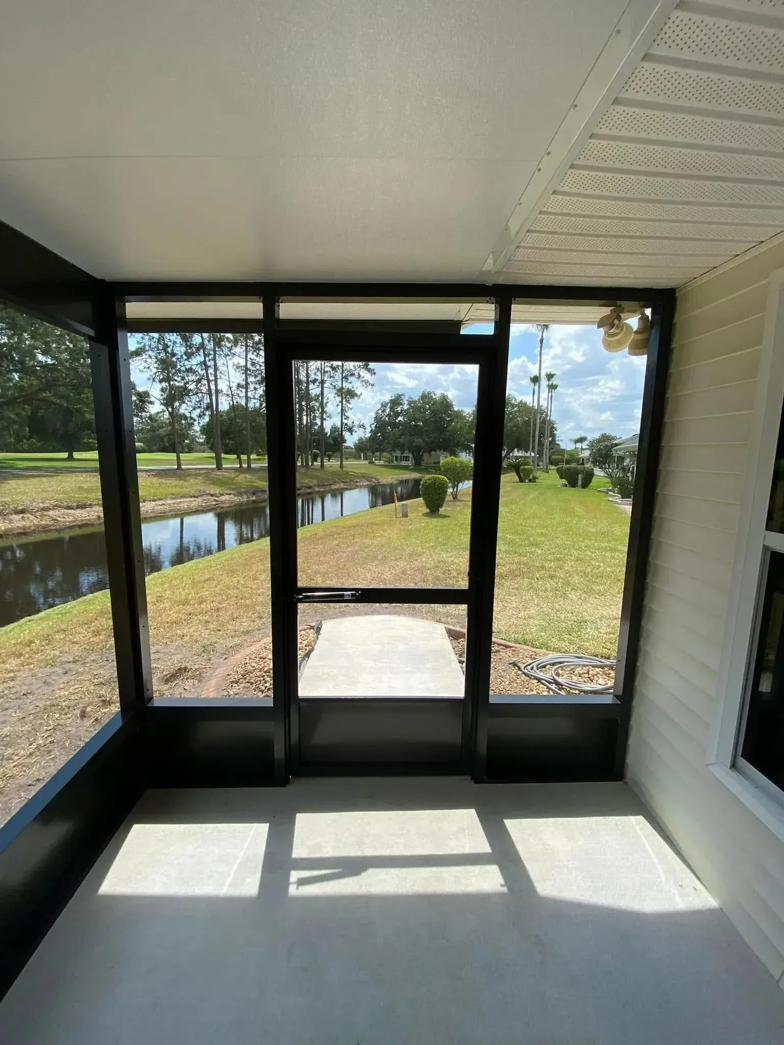 Sunlit screened porch with white ceiling, black-framed doors, and view of canal and grassy yard