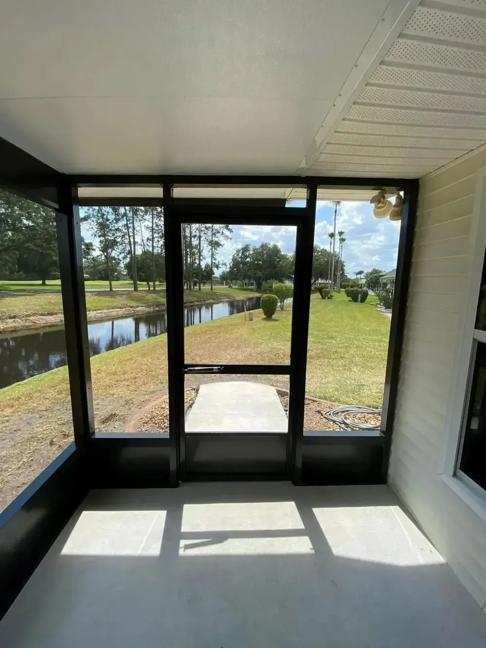 Sunlit screened porch with white ceiling, black-framed doors, and view of canal and grassy yard