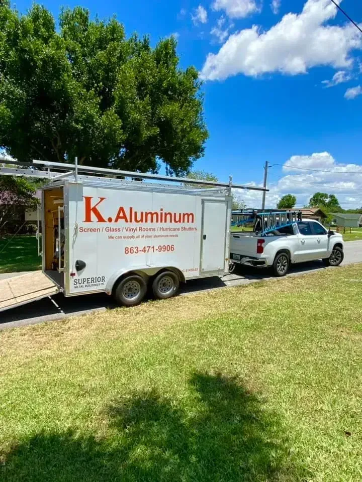 White utility trailer and truck parked on a sunny grassy lawn, with a large green tree behind them.