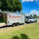 White K. Aluminum trailer hitched to a silver SUV on a sunny grassy roadside.