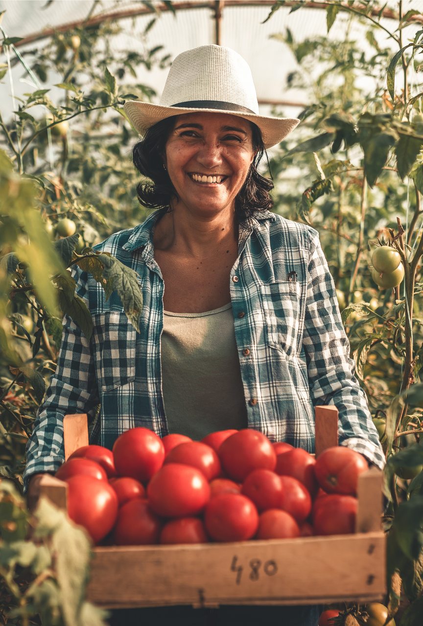 Woman in a straw hat smiles, holding a wooden crate of ripe red tomatoes in a greenhouse.