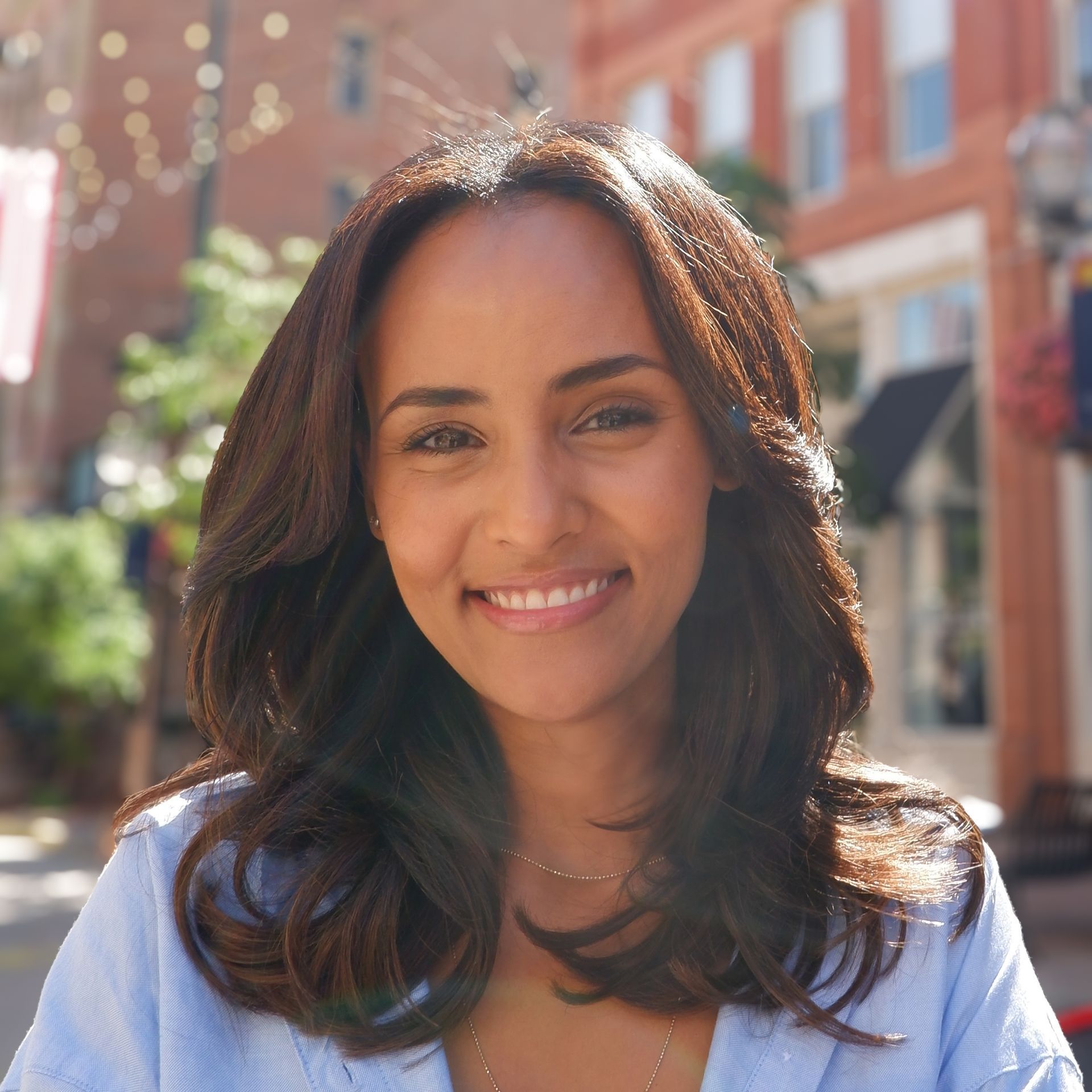 Woman with brown hair smiling outdoors, downtown street in background.