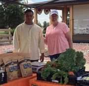 Two people at a farm stand with produce and bags. Man wears yellow shirt, woman pink shirt.