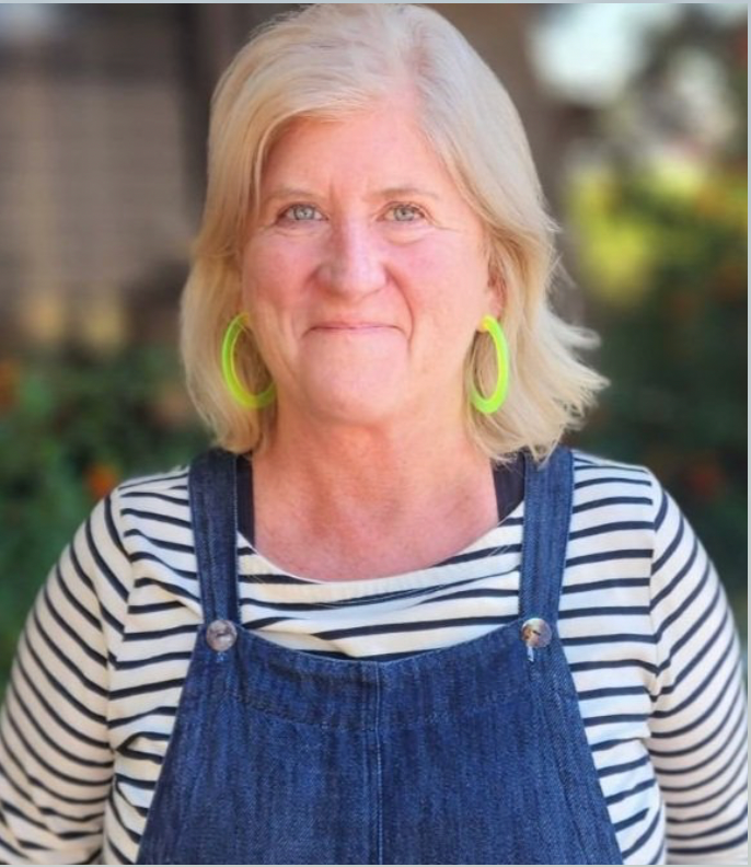 Woman with blonde hair wearing striped shirt, denim overalls, and neon green earrings, smiling.