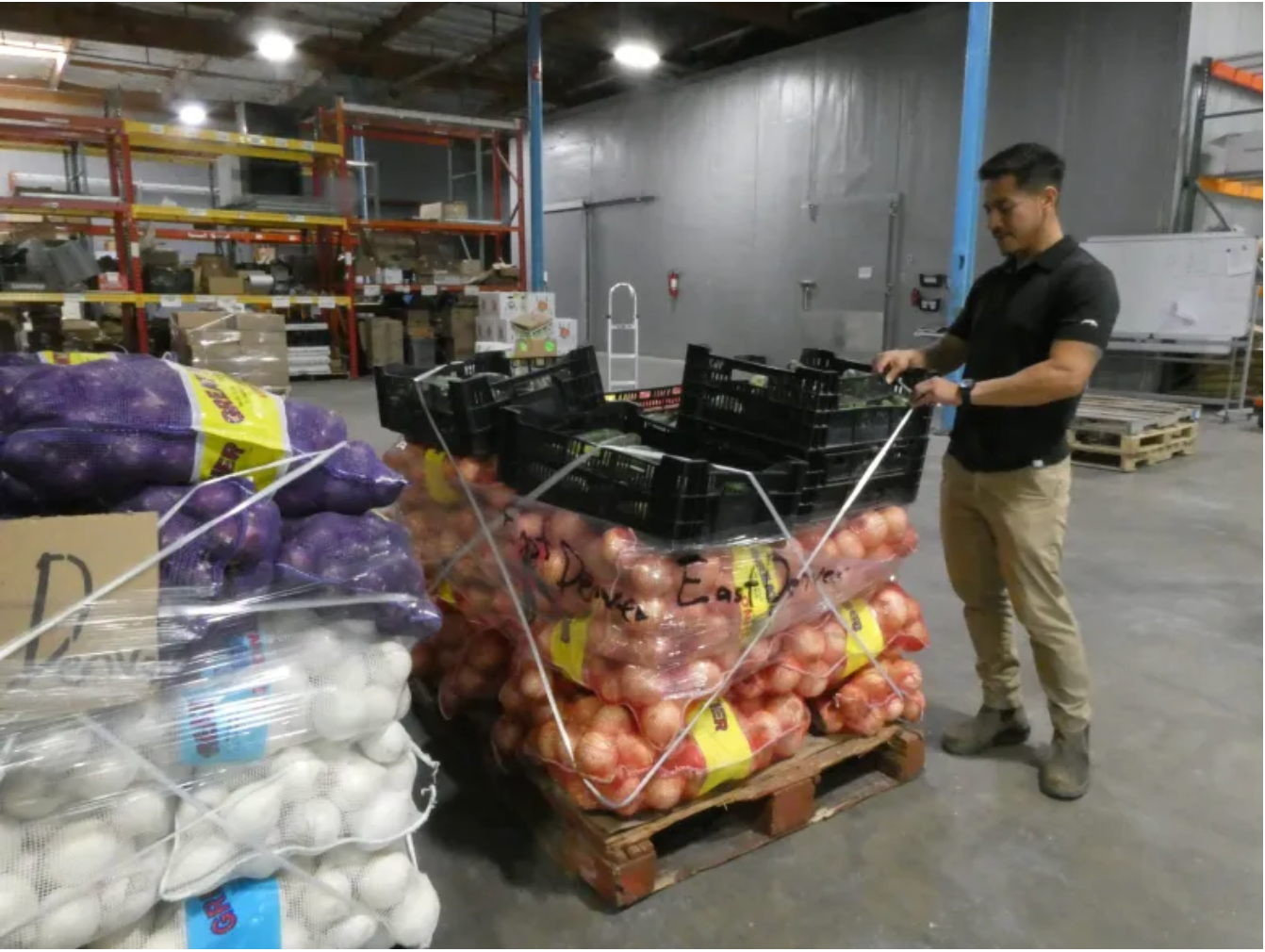 Man in a warehouse pulling a pallet of onions and empty black crates. Other onion bags are on pallets.
