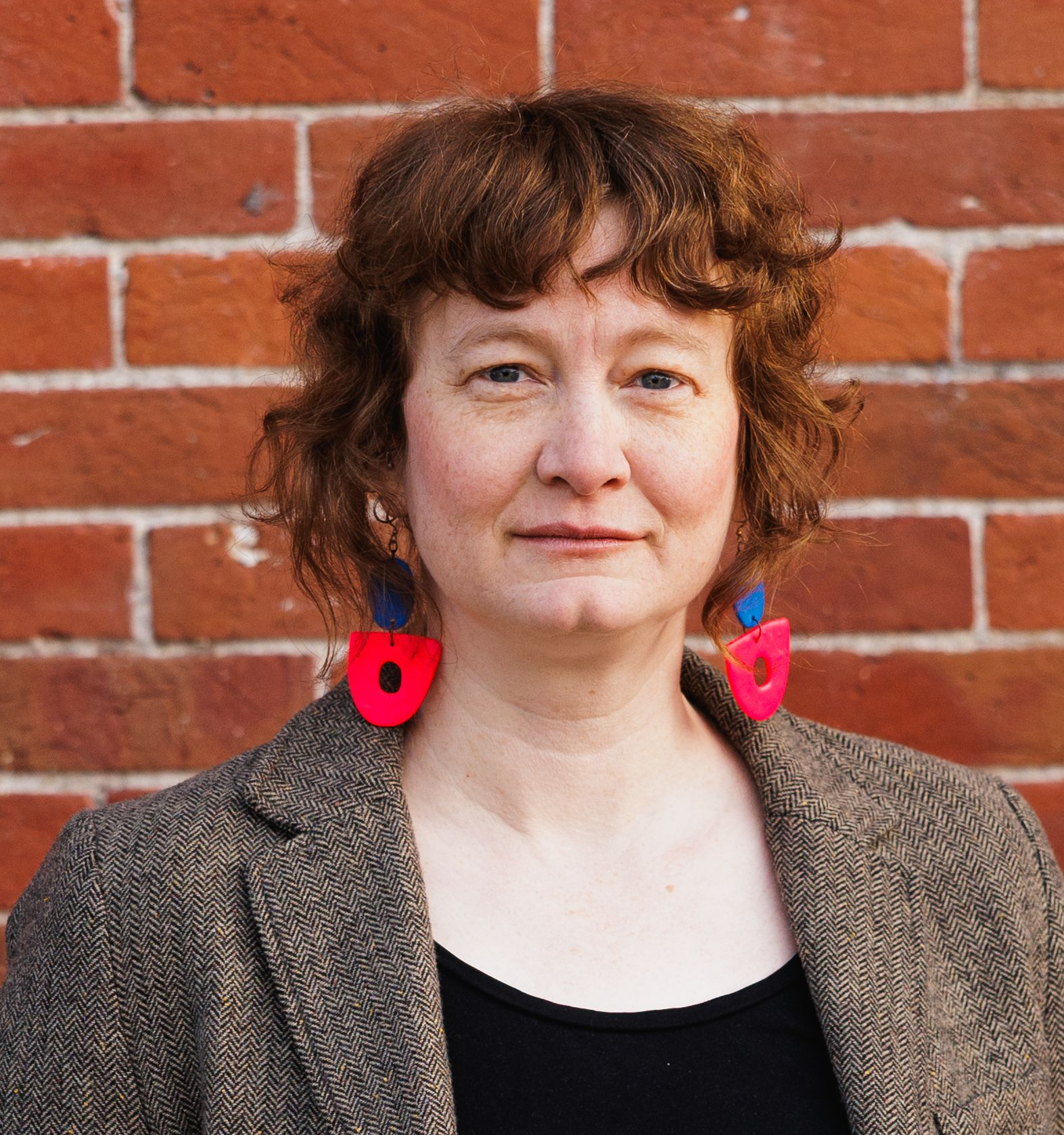 Woman with red hair, wearing a black shirt and gray blazer, in front of a brick wall.