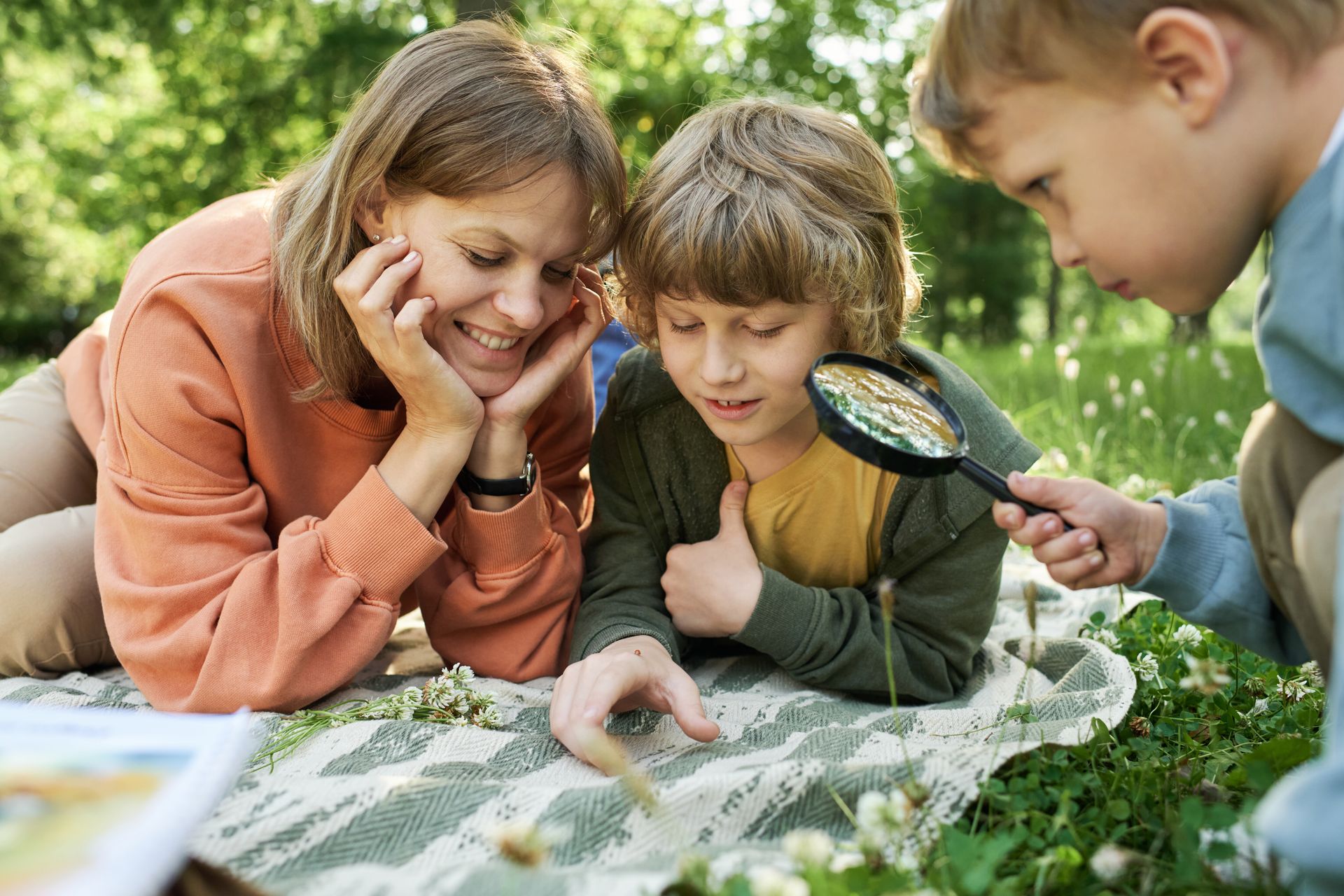 A woman and two children lie on a blanket outdoors, using a magnifying glass to examine something on the ground.