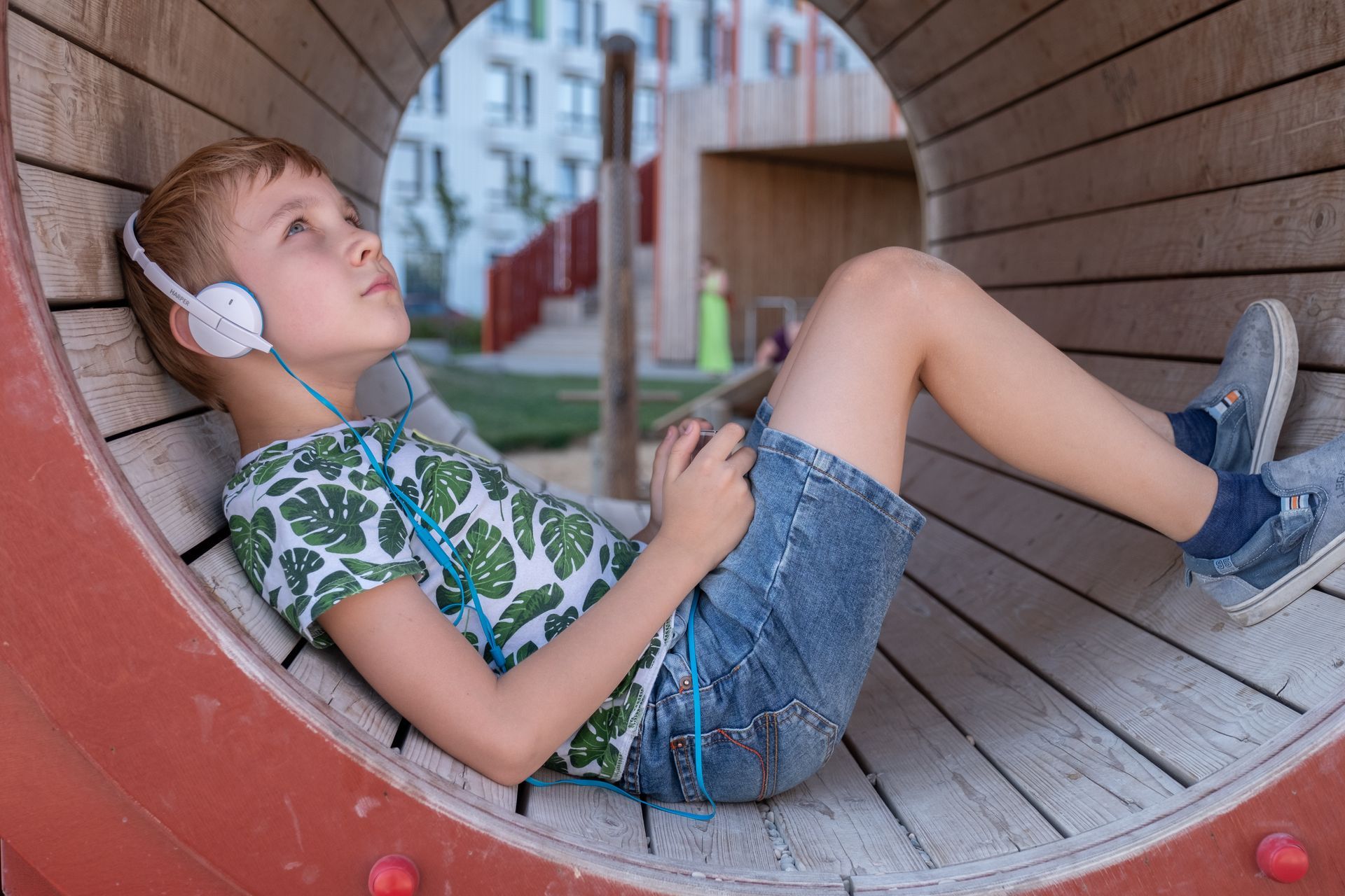 A child wears white headphones while lying inside a wooden circular tunnel structure, looking upward thoughtfully.