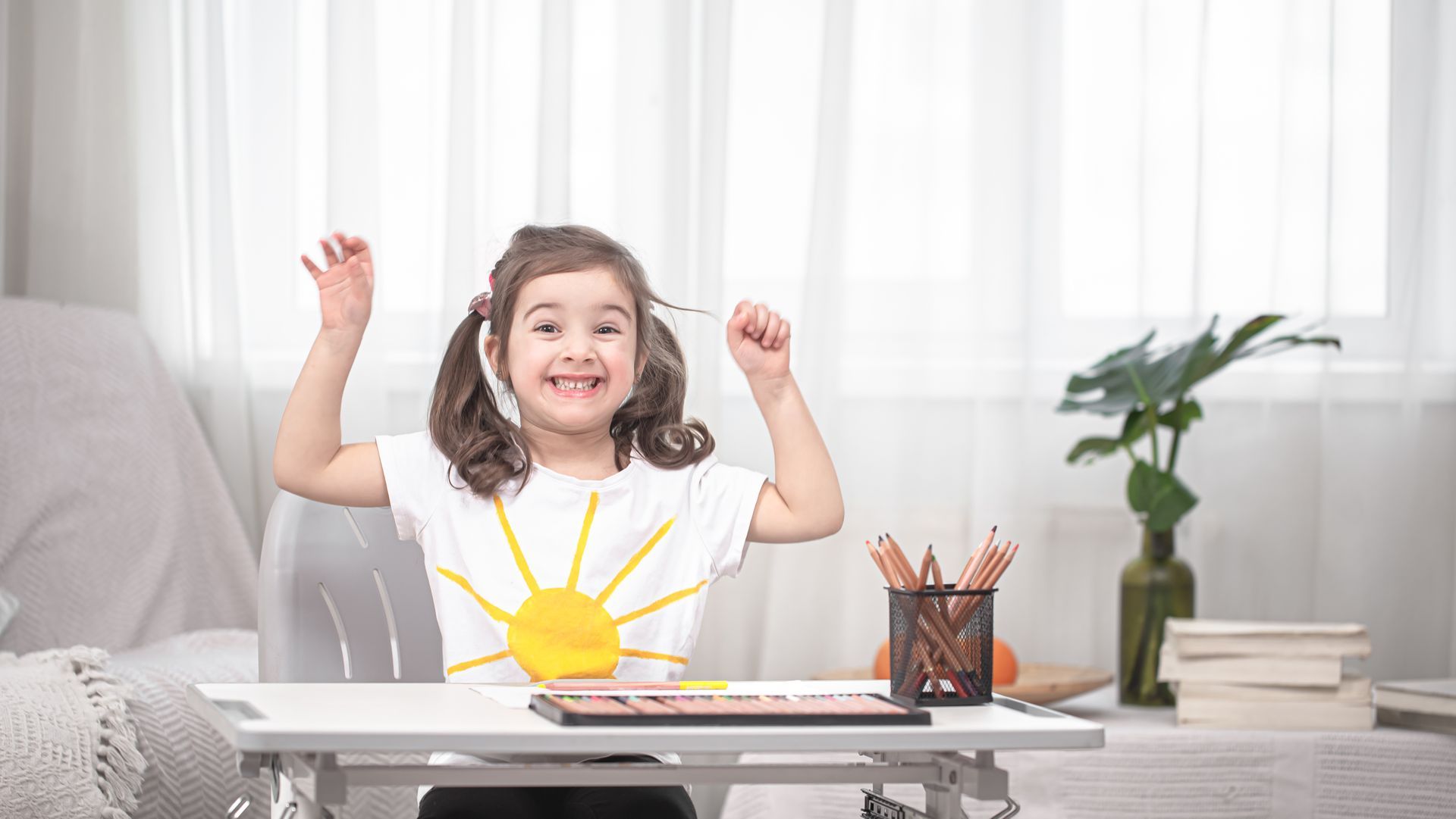 A smiling child wearing a sun-themed shirt sits at a desk, raising both hands excitedly in a bright, indoor setting.