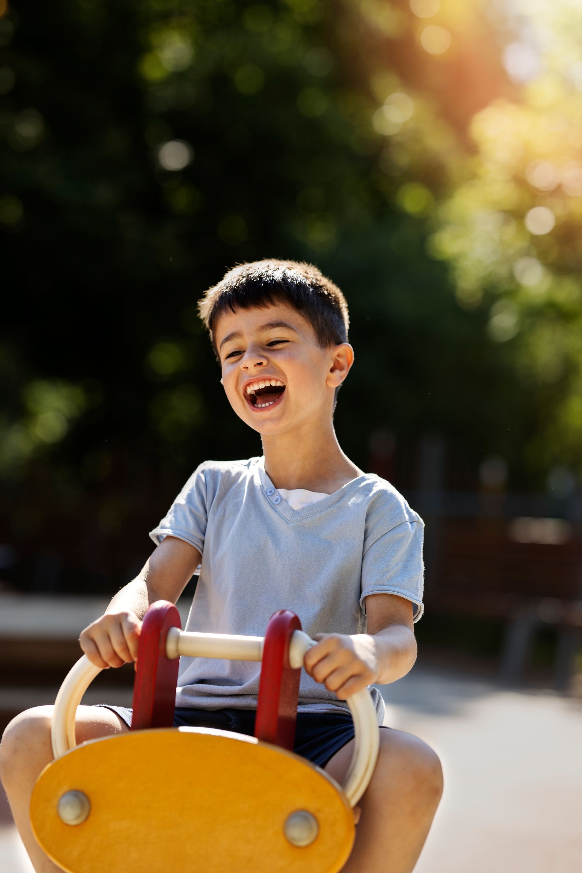 A joyful child laughing while riding a bright yellow playground rocker in a sunny, tree-filled park.