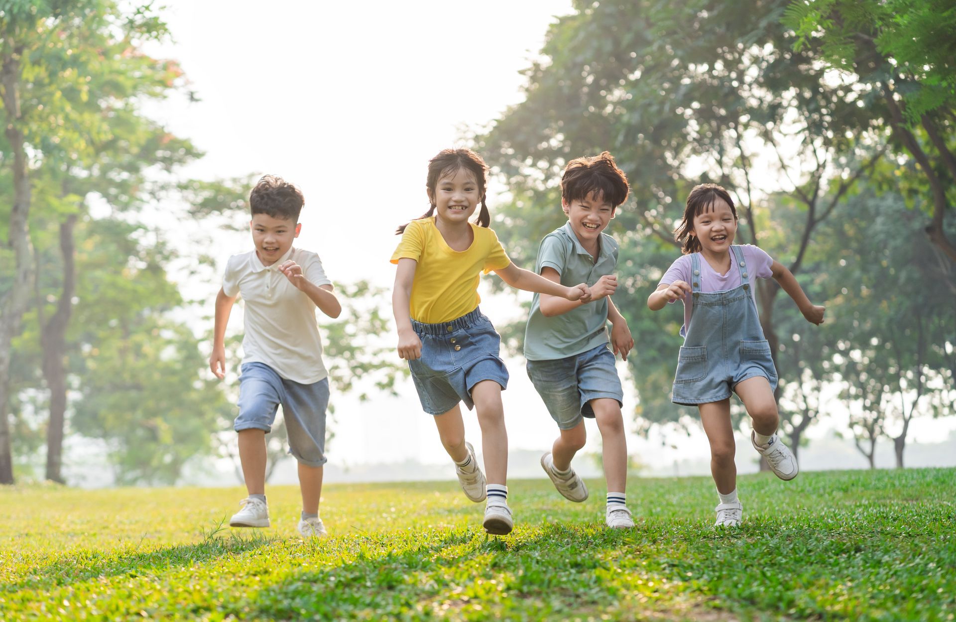 Four children run joyfully across a grassy park, their faces lit with smiles in the warm sunlight.