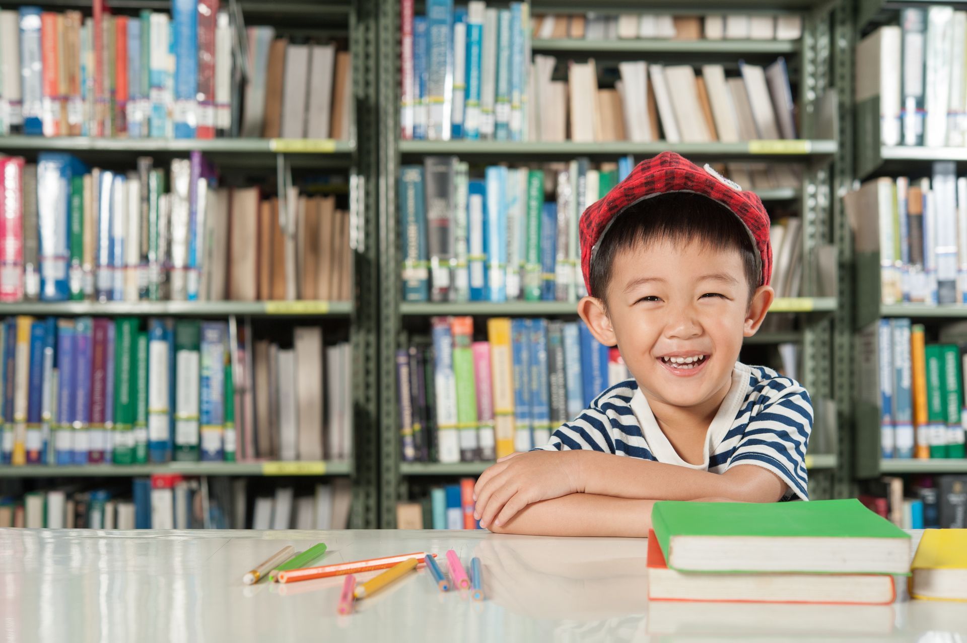A smiling child in a red cap sits at a table with books and pens in a library with bookshelves in the background.
