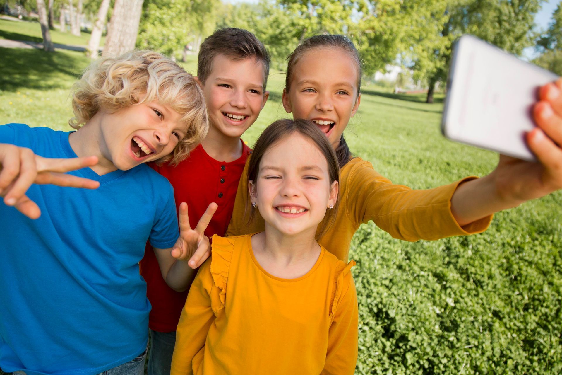 Four children in colorful shirts laugh while taking a selfie together in a sunlit grassy park.