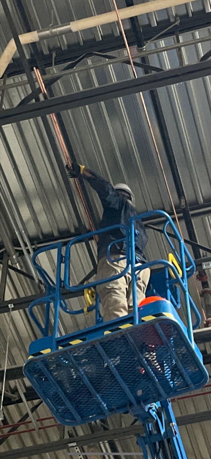 Worker in a blue elevated lift near a corrugated metal ceiling, reaching up to inspect or repair overhead beams.