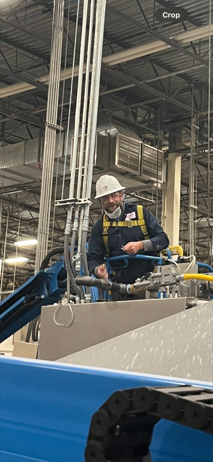 Worker in a hard hat standing on equipment inside an industrial warehouse with overhead pipes and beams