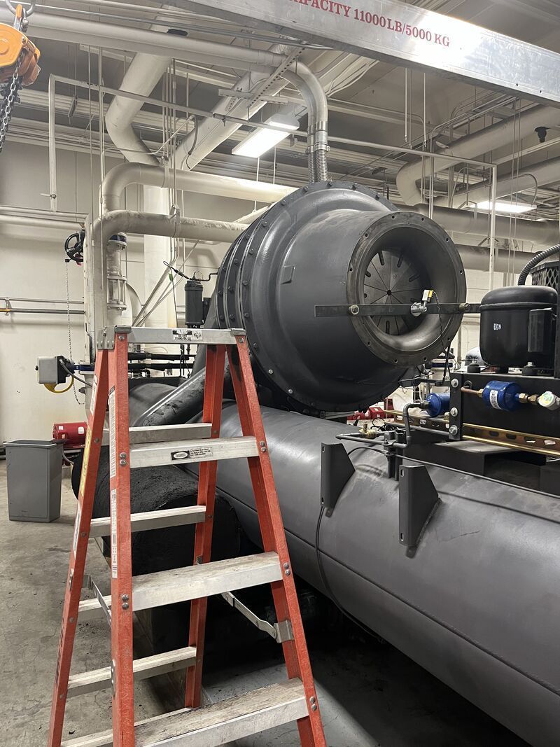 Industrial HVAC equipment in a utility room, with a ladder beside a large black duct and machinery.