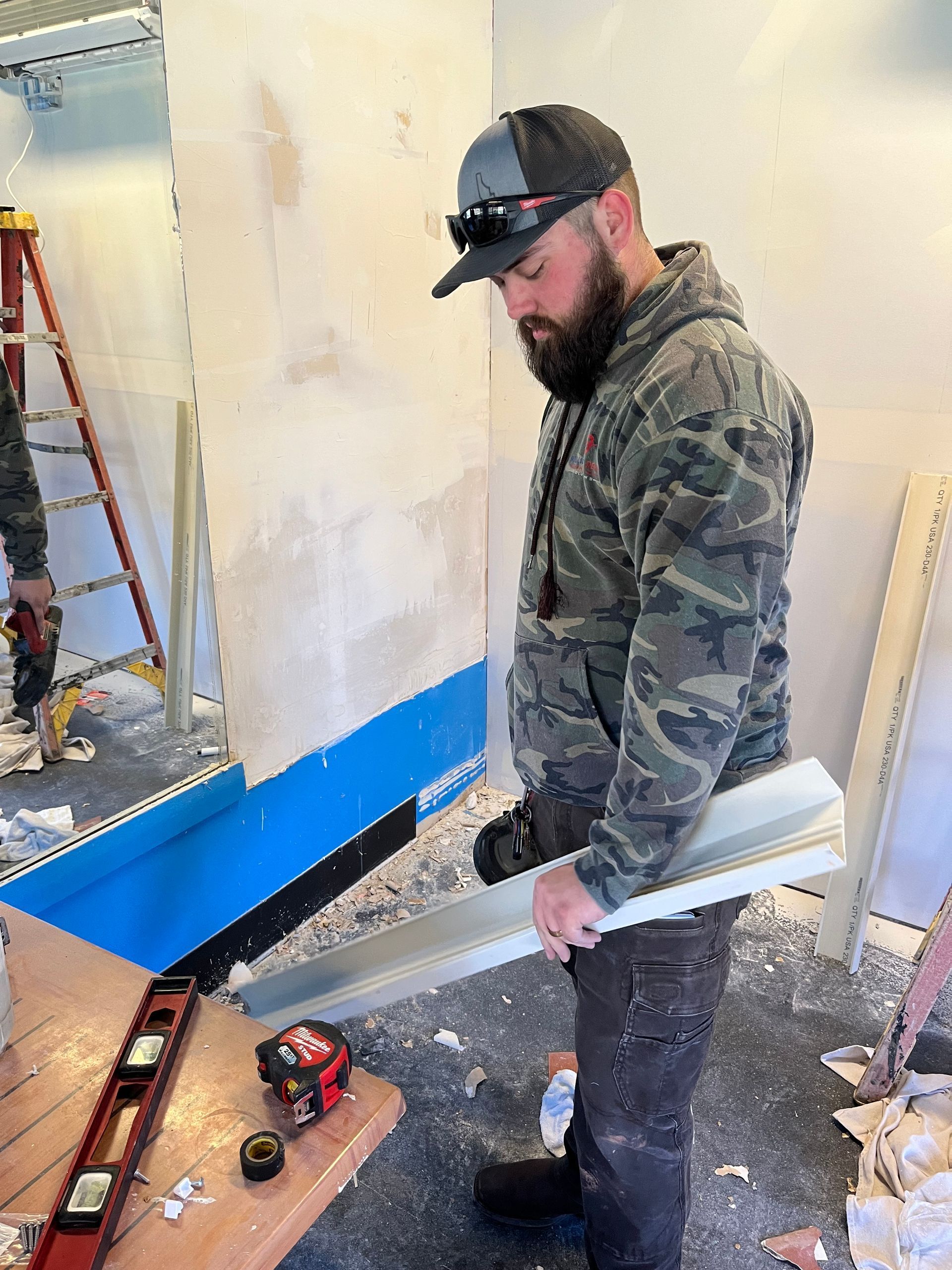 Worker in camouflage hoodie and cap stands in a partially renovated room with tools and debris.