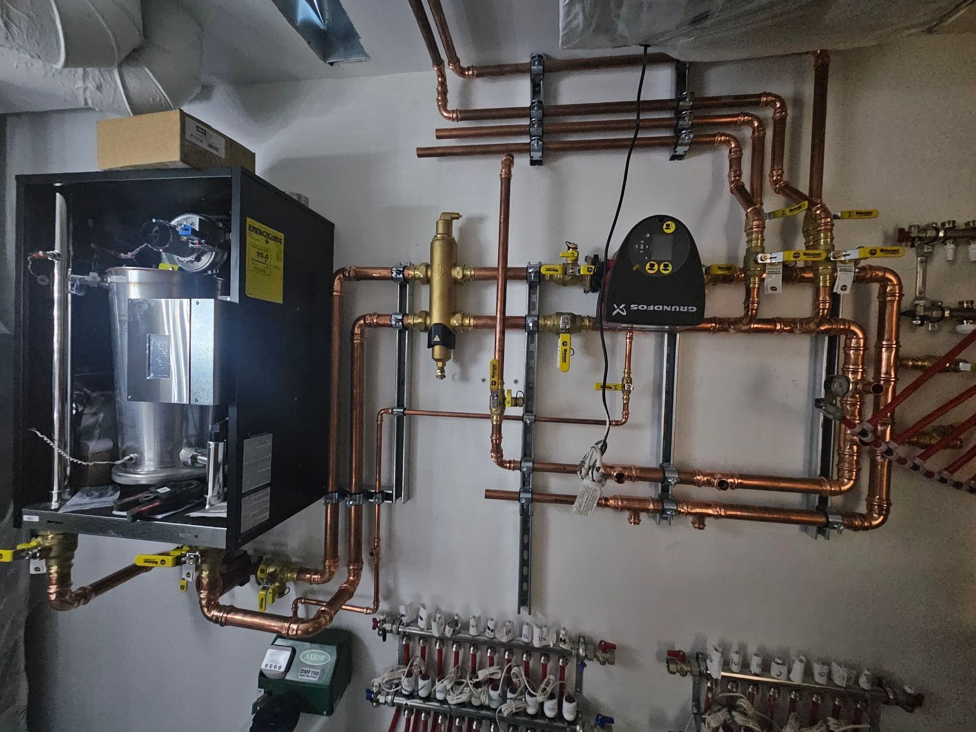 Boiler room with wall-mounted copper pipes, valves, and a large black heating unit on the left.