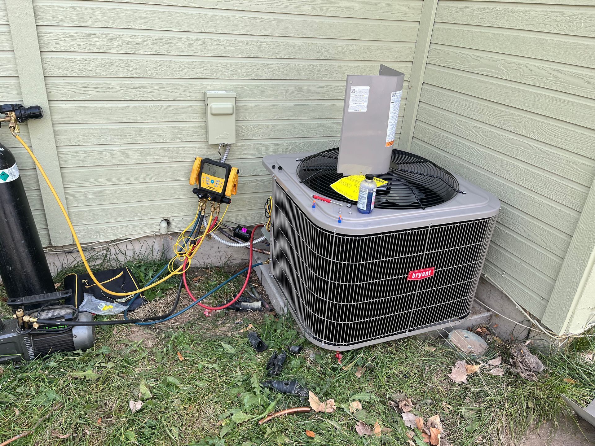 Outdoor HVAC condenser unit beside a house wall with gauges and hoses attached for servicing
