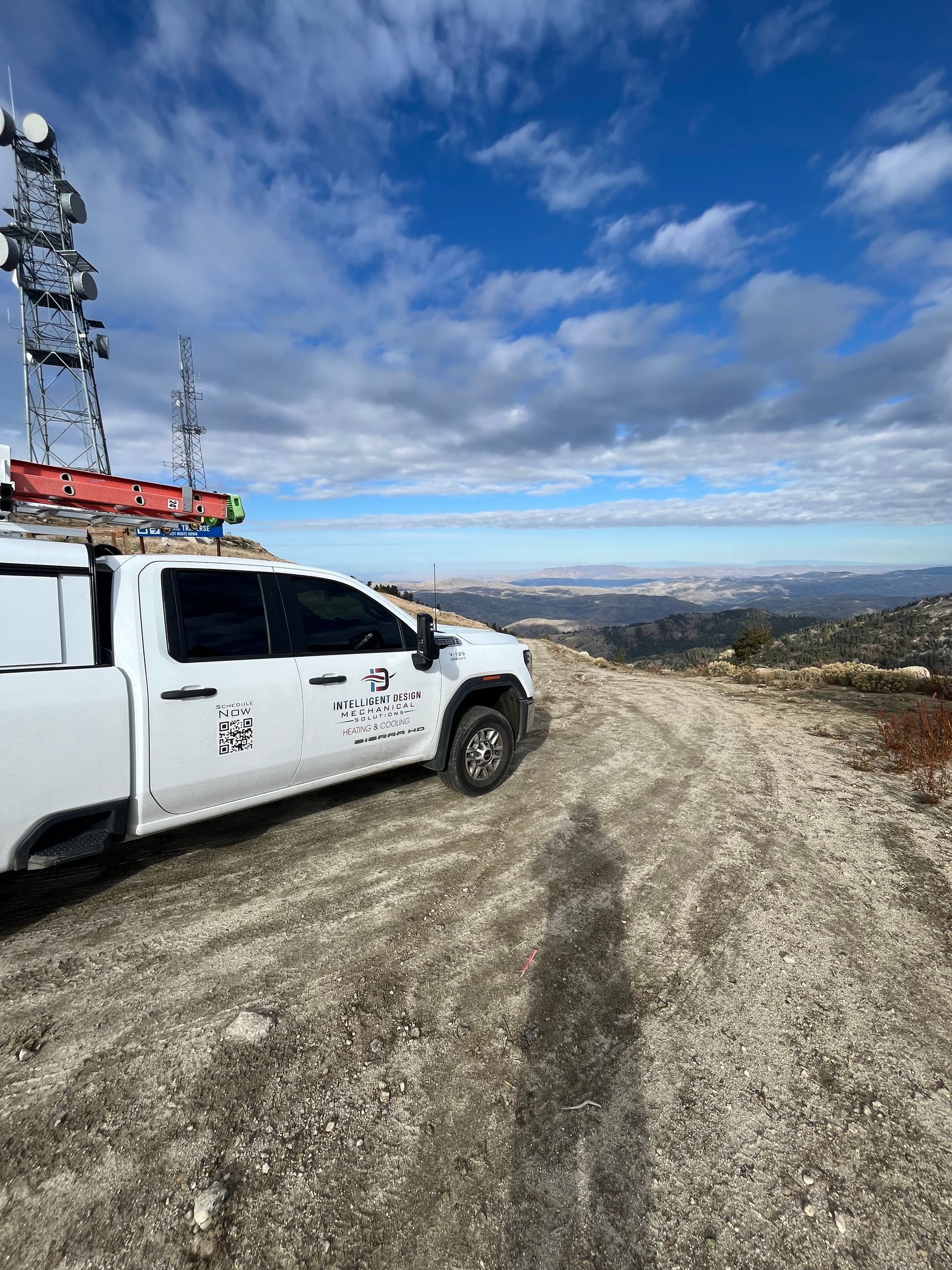 White utility truck on a rocky hilltop with a radio tower, overlooking a distant landscape under a cloudy blue sky