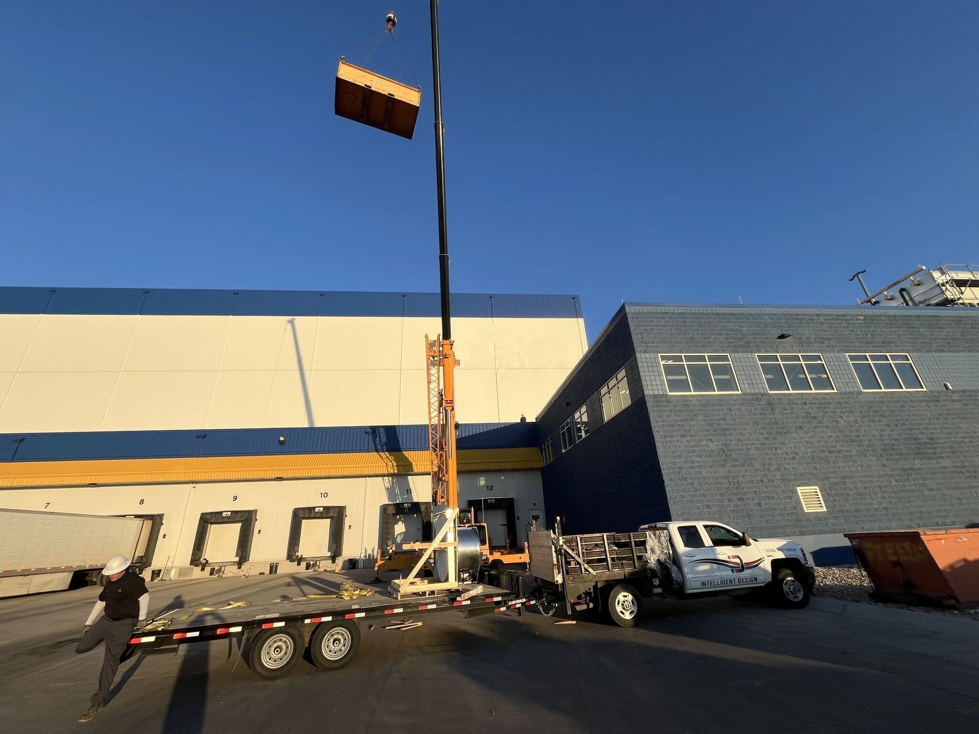 Workers use a crane to lift a large orange object beside a blue industrial building and truck.