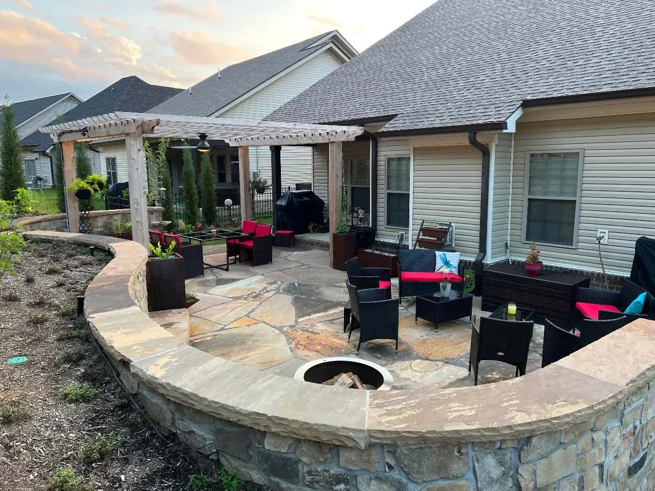 Curved stone patio with black outdoor chairs and red cushions beside a beige house.
