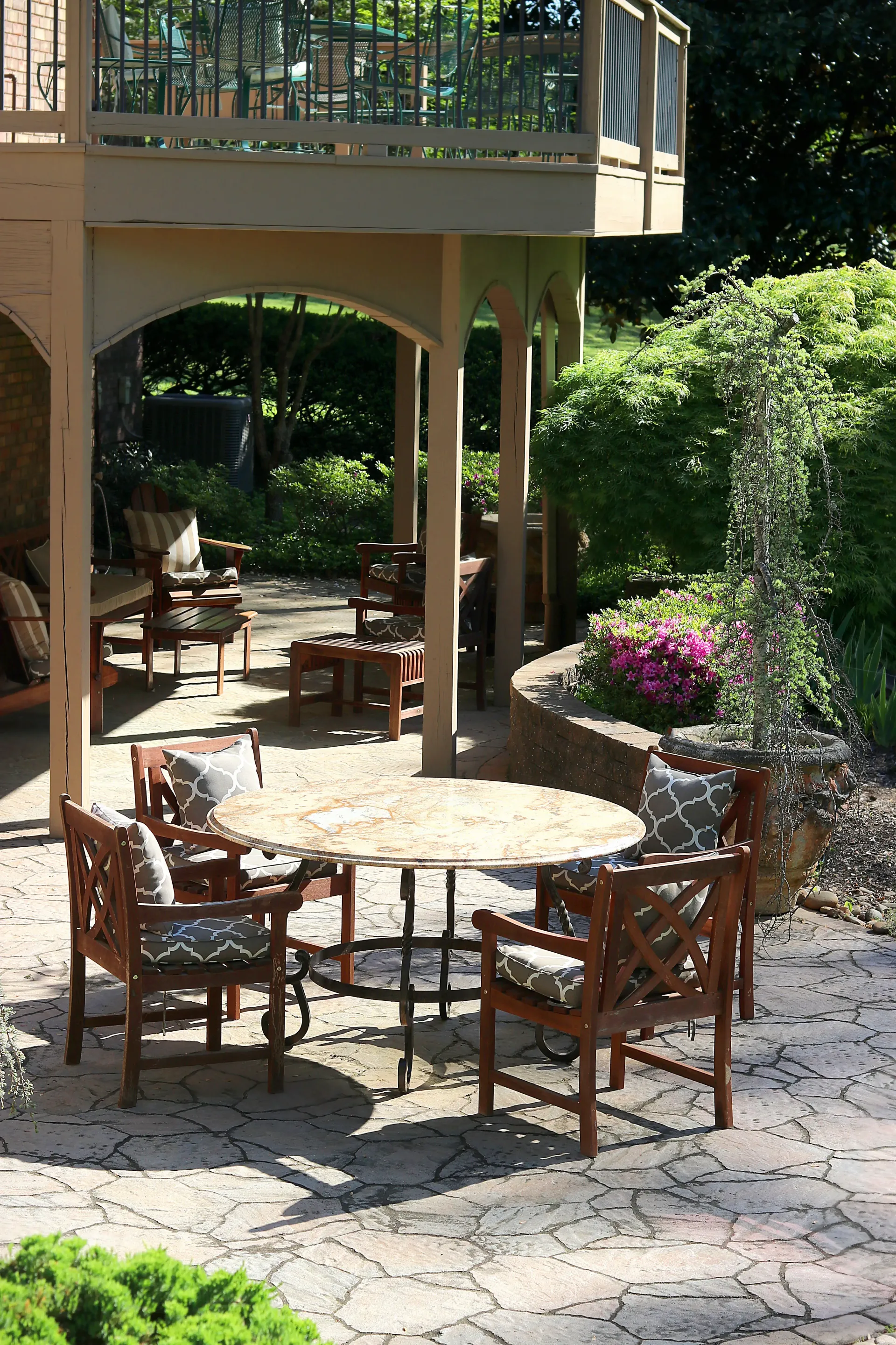 Wooden patio chairs around a round table on a sunny stone terrace beside a garden and deck