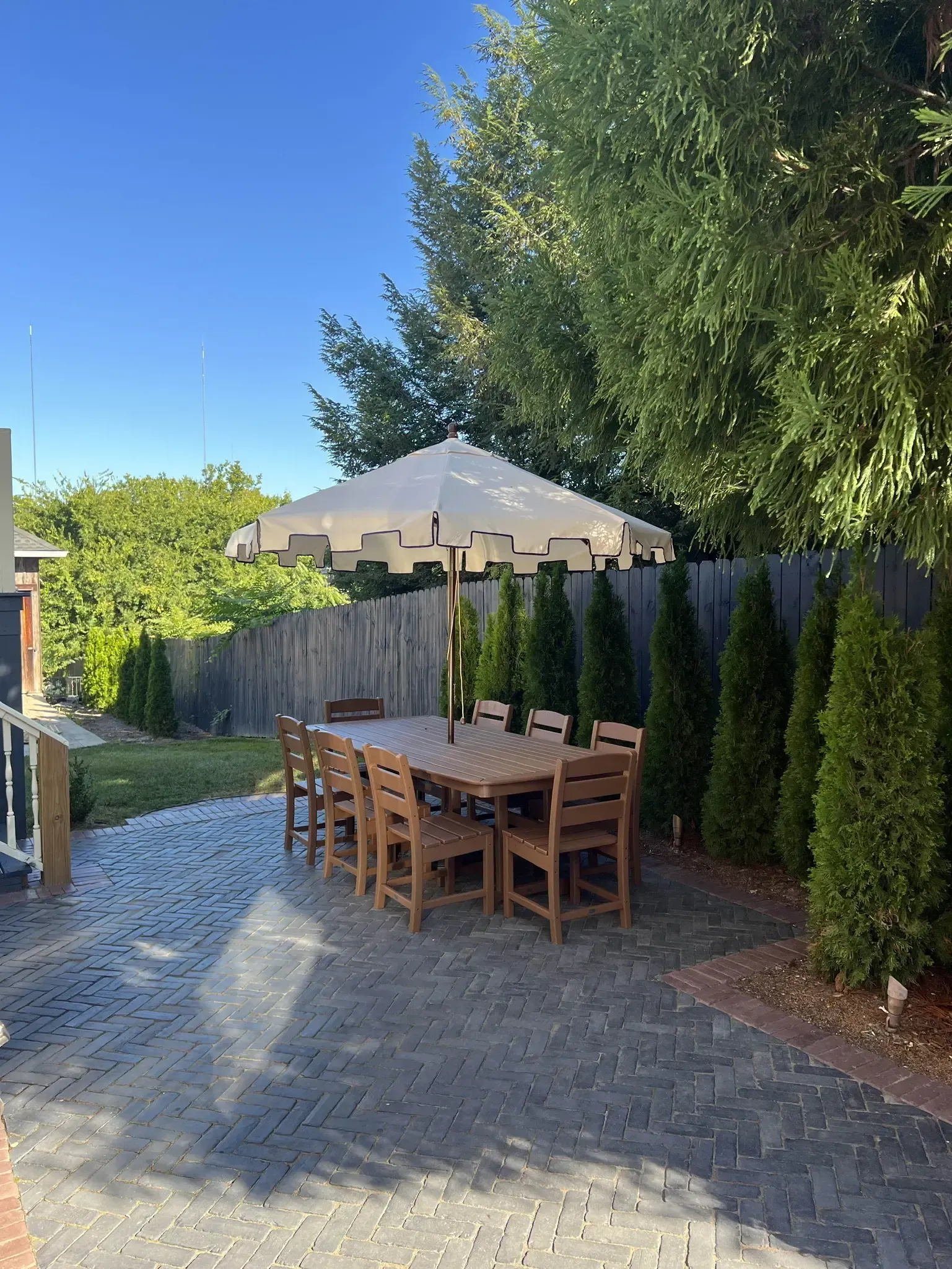 Patio with wooden dining table and chairs beside a hedge and fence under a clear blue sky
