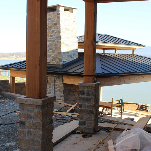 Covered outdoor patio under construction with wooden posts, stone columns, and a metal roof.