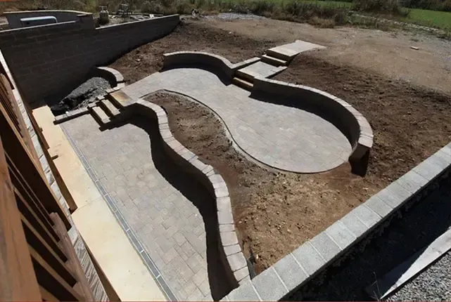 Empty concrete pool layout under construction in a dirt yard, with curved basin walls and steps.
