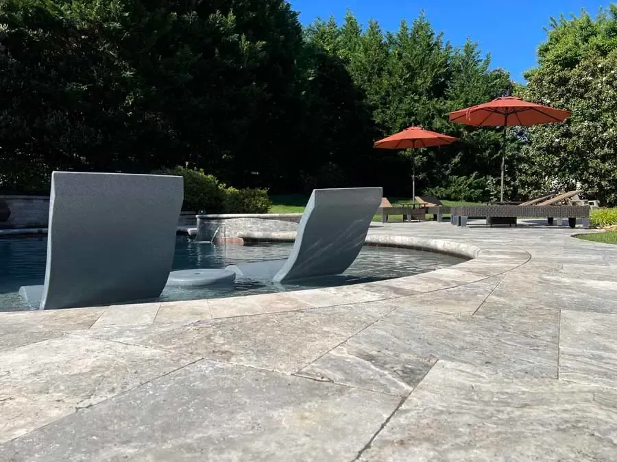 Modern backyard pool with curved silver lounge chairs, stone patio, and red umbrellas near trees
