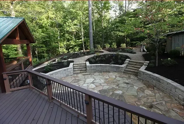 Stone patio with curved seating walls and wooden deck in a wooded backyard.