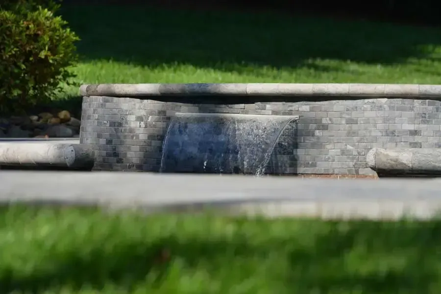 Stone fountain wall with a small waterfall in a landscaped garden, framed by blurred grass in the foreground