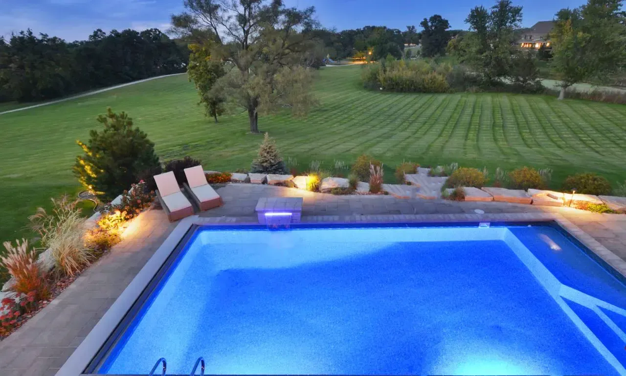 Lit backyard pool overlooking a green lawn at dusk, with lounge chairs and warm landscape lighting