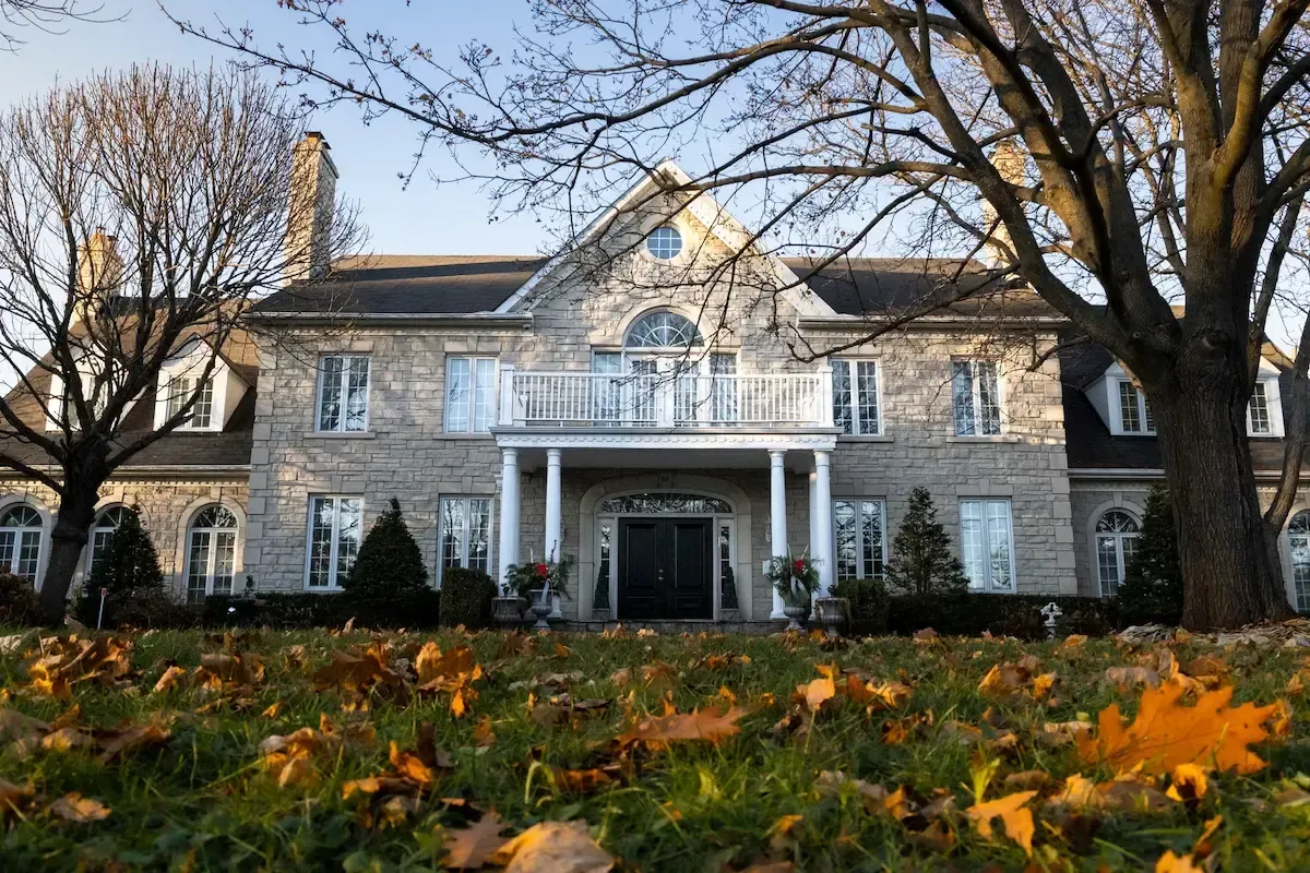 Large stone house with white columns and balcony, framed by bare trees and autumn leaves in front.