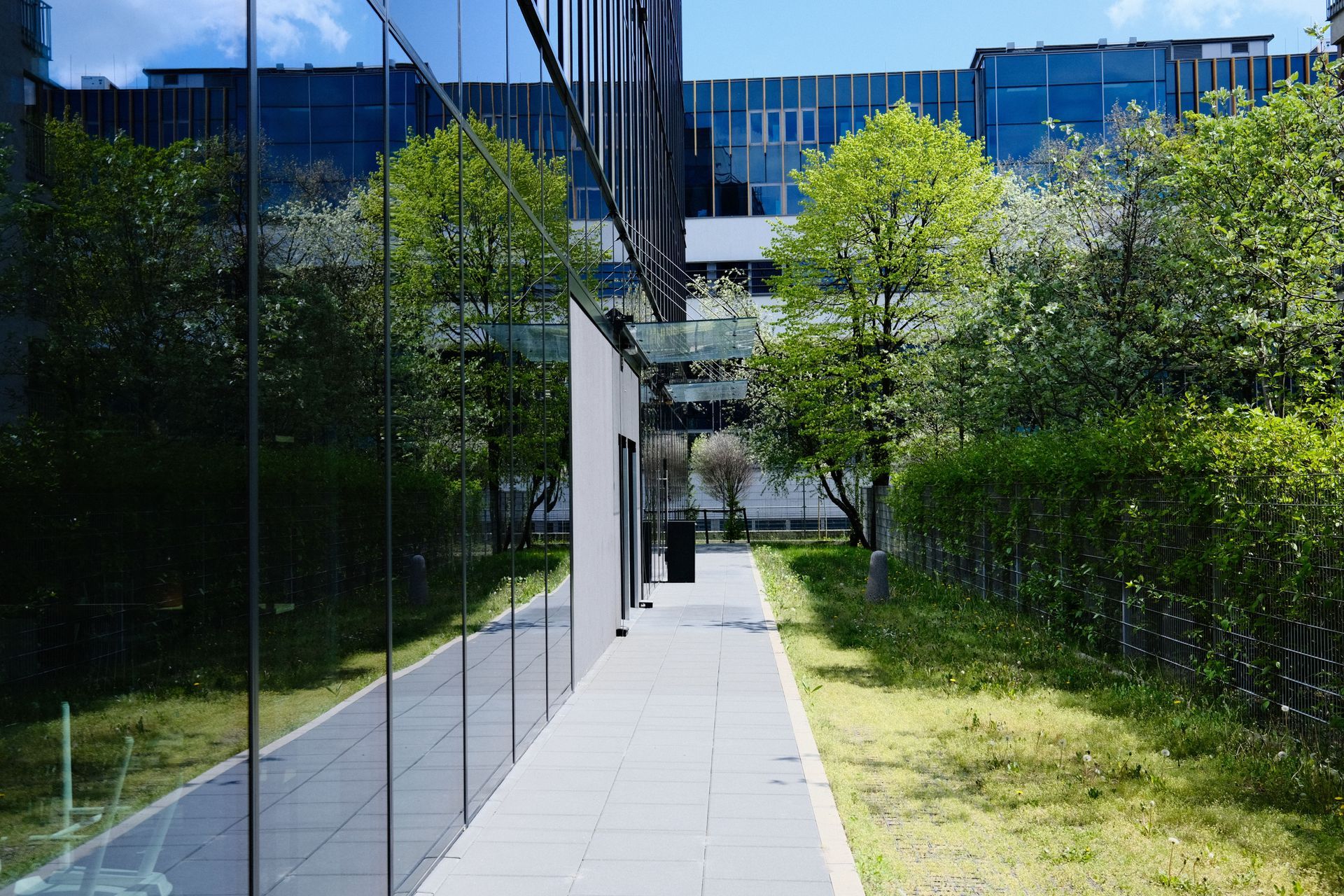 Modern office building walkway with glass walls, green trees, and landscaped grass under blue sky