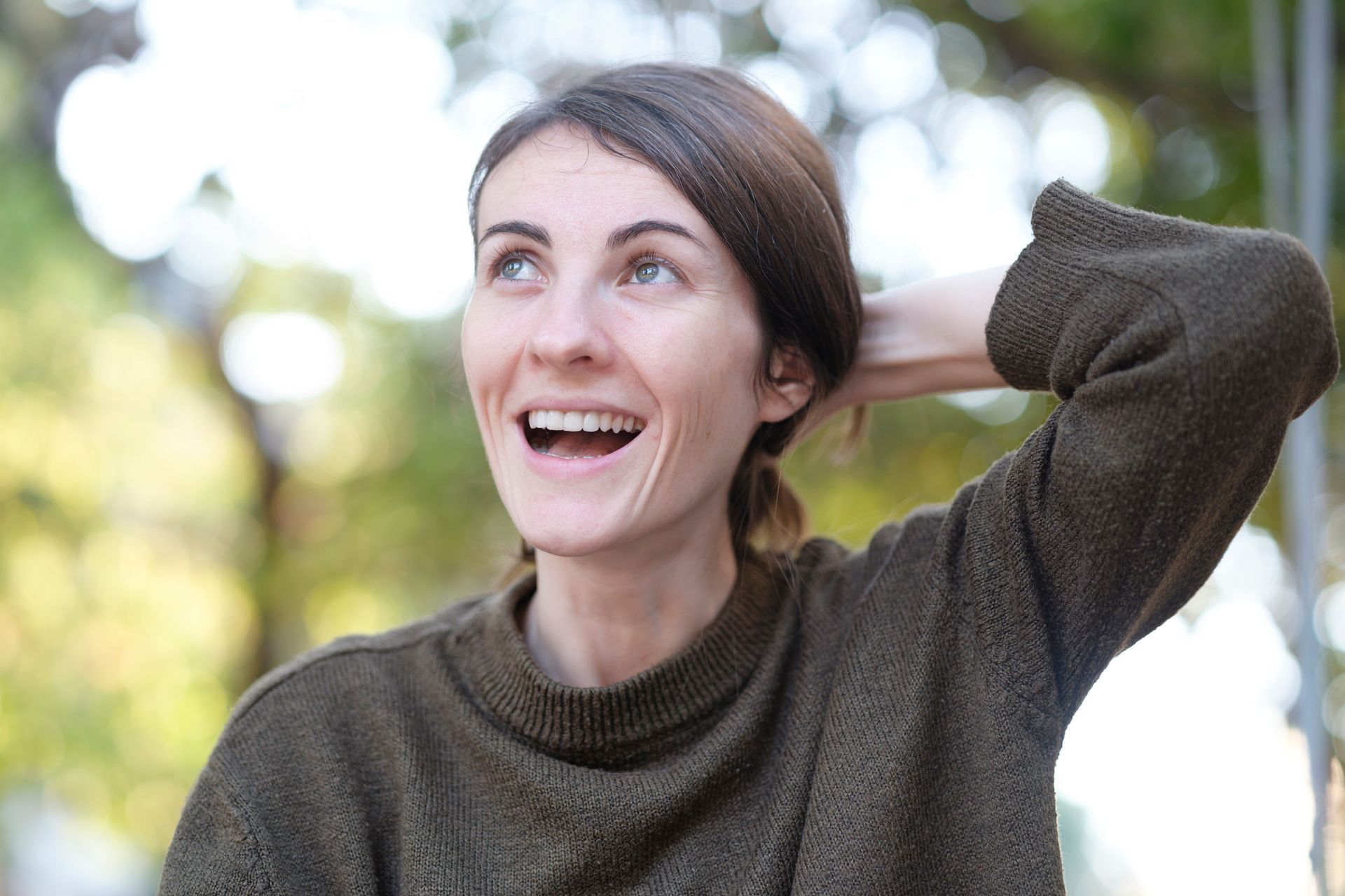 Smiling person in a green sweater outdoors with hand behind head