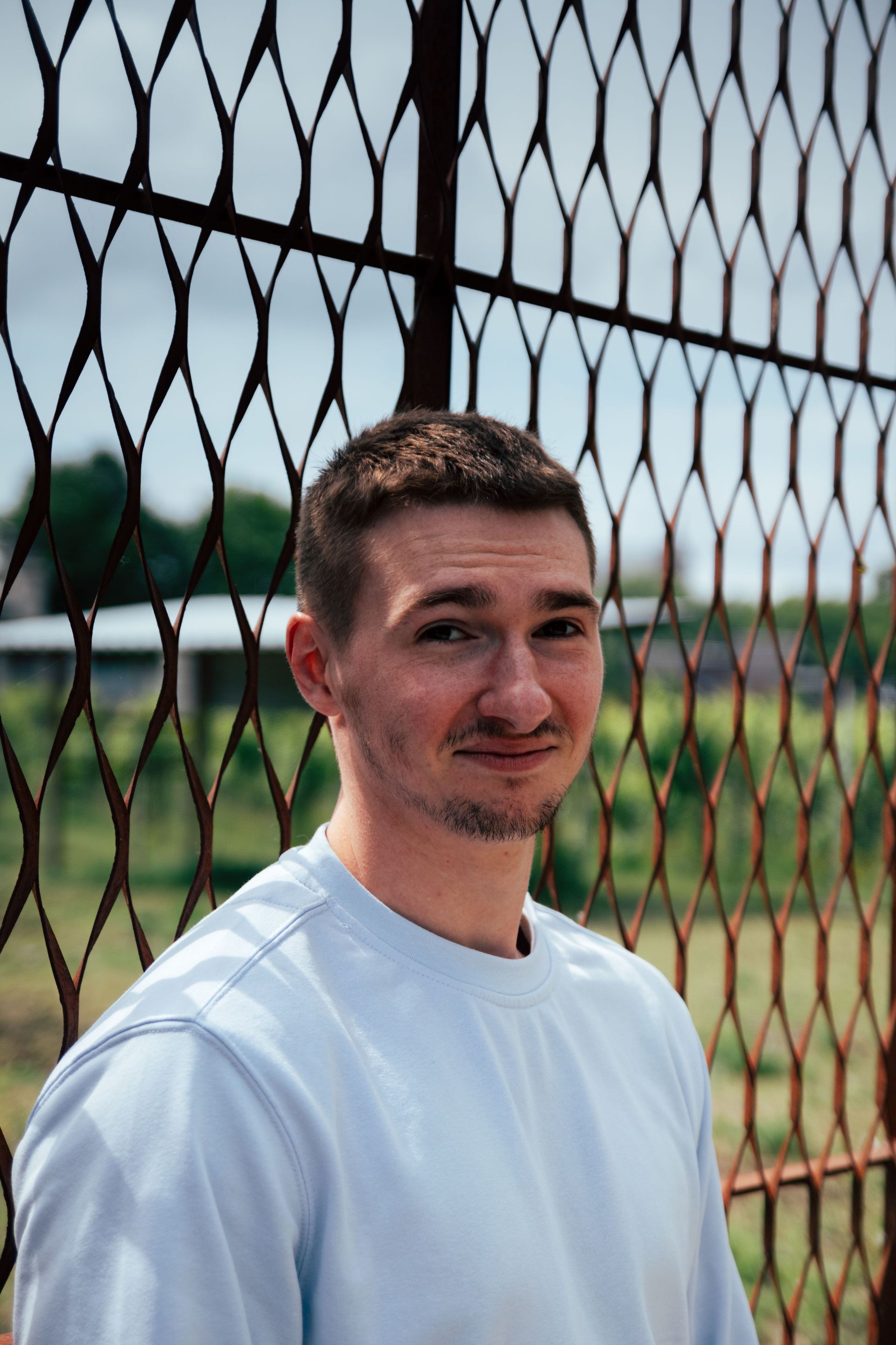 Person in a white shirt standing by a chain-link fence outdoors, with a baseball field in the background.