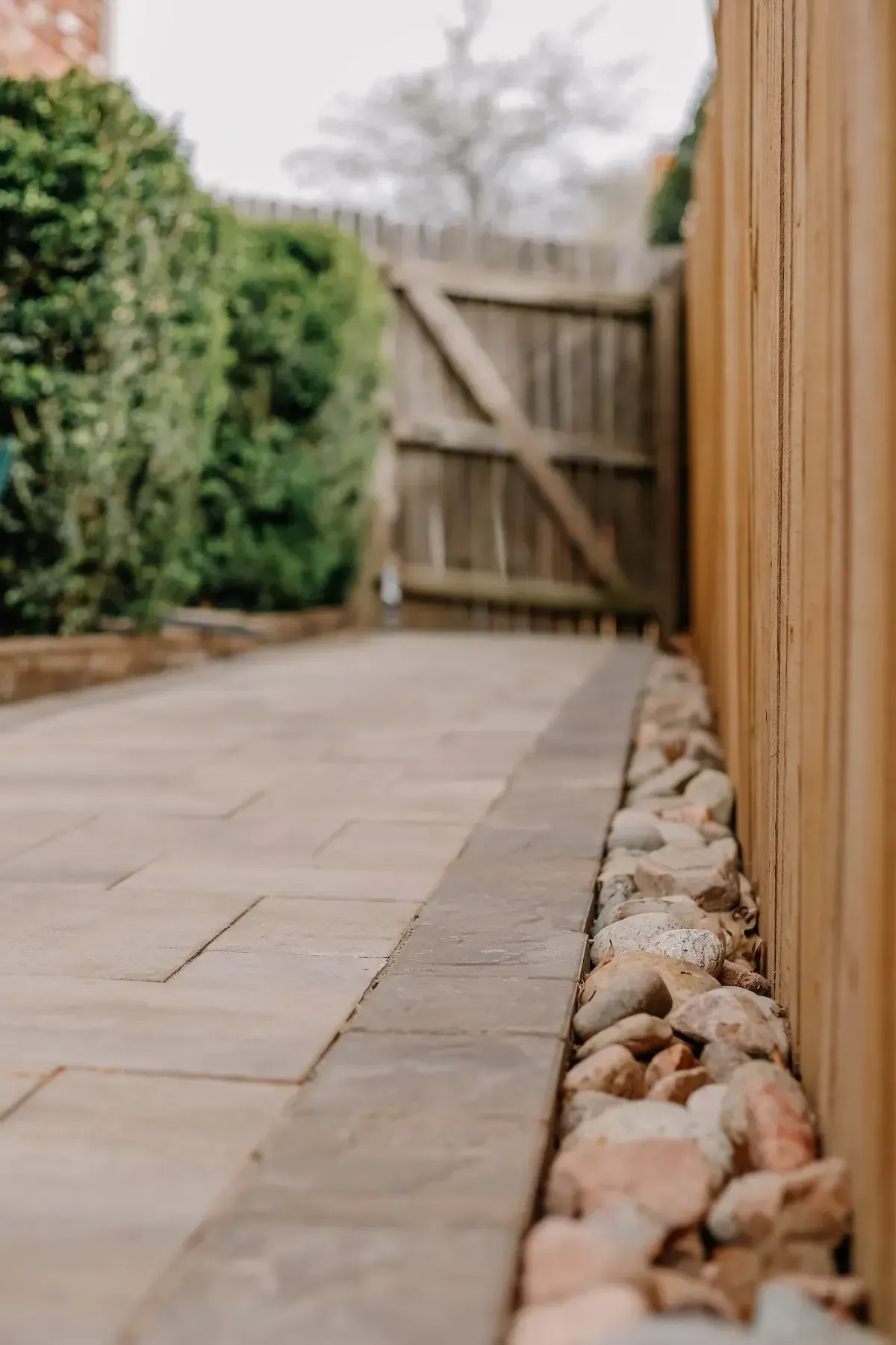 Stone patio beside a wooden fence, with pebbles lining the edge and a gate in the background.