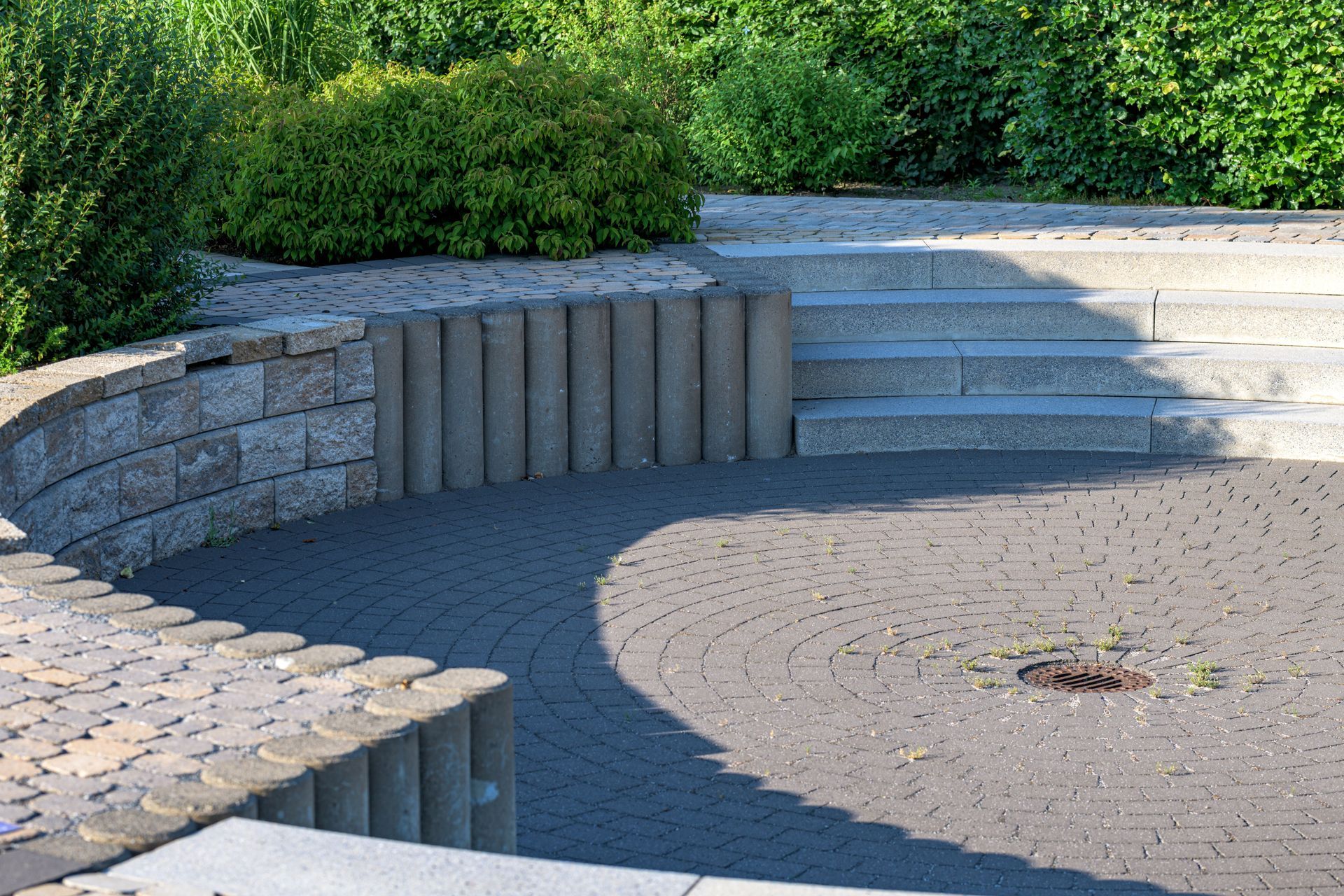 Curved stone seating around a circular paved courtyard with a small fire pit, bordered by green shrubs