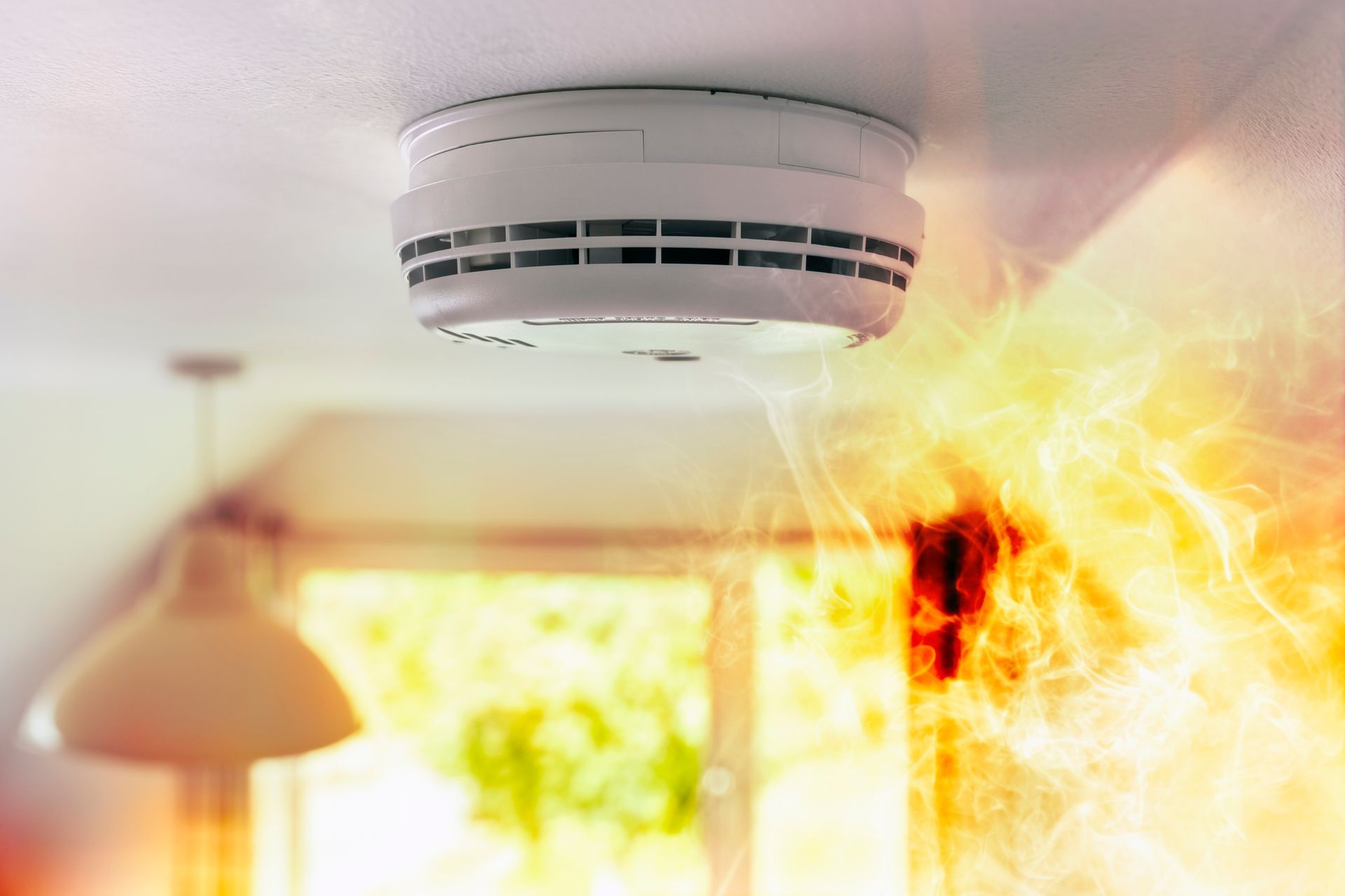 A smoke detector is hanging from the ceiling in a room with smoke and flames around it
