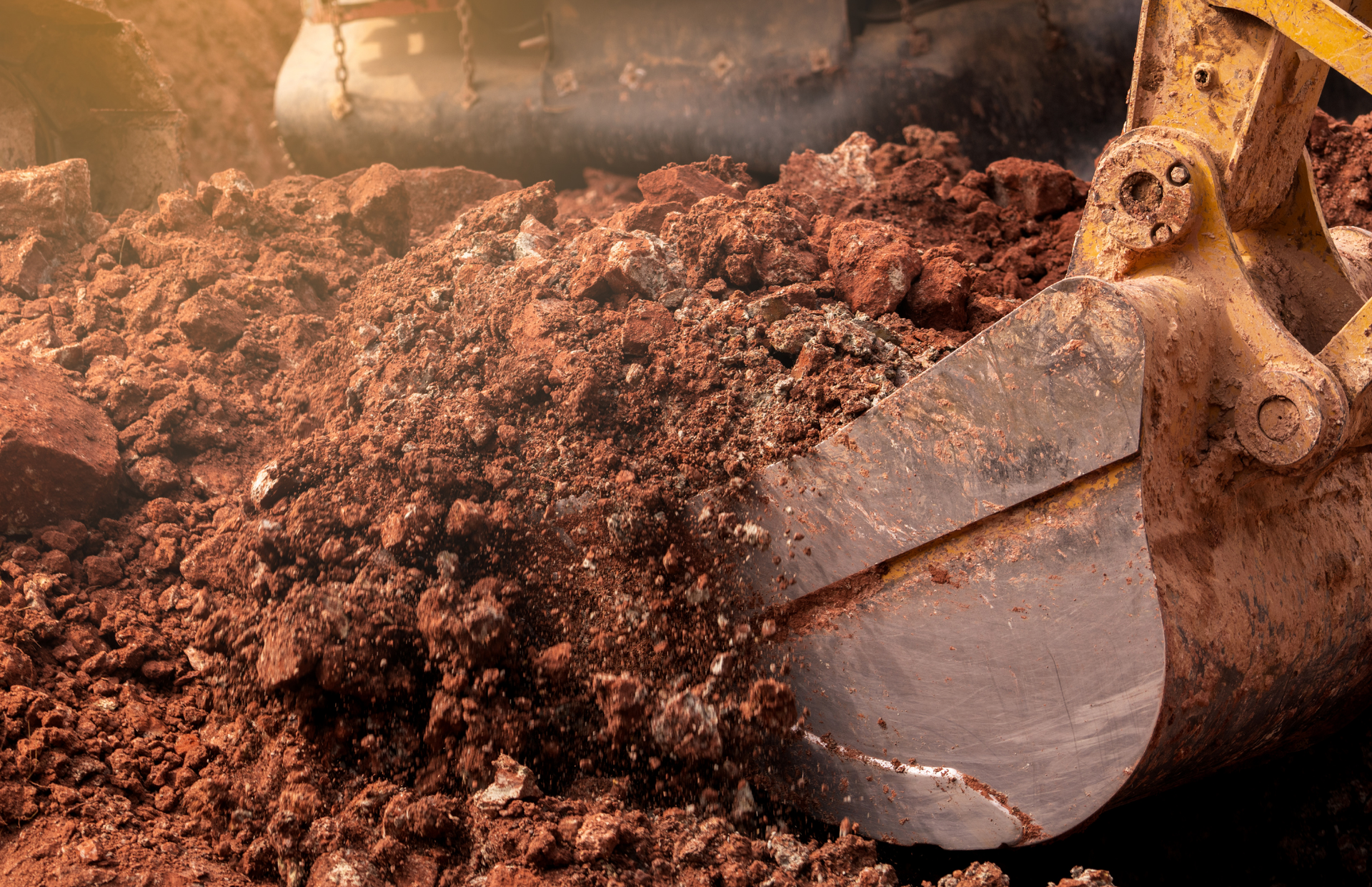 Excavator bucket scooping up reddish-brown dirt and rock at a construction site.