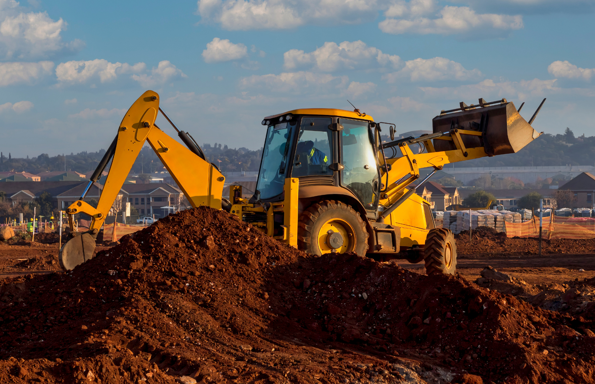 Yellow backhoe on a dirt pile, digging at a construction site with buildings in the background.